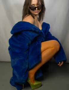 A young woman crouching down and posing with sunglasses and wearing a bright blue fur coat and neon green heels.