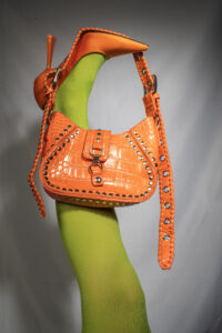 A leg in a green nylon and an orange high heel balancing an orange purse for a fashion photoshoot.