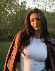 A woman posing in a forest with the sunset shining on her face, she is wearing a white body suit and a maroon leather jacket is draped over her shoulders.