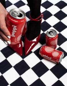 Someone holding a classic cola Poppi can while standing on a black and white checkered floor, two other cans resting beside the shoes.