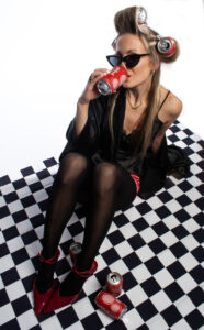 A young woman with Poppi cans as rollers in her hair sitting on a checkered floor drinking a classic cola Poppi.