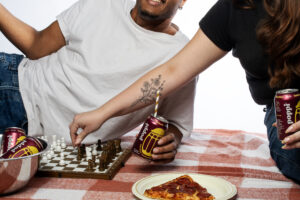 Two friends enjoying pizza and sipping on Poppi rootbeer cans while playing chess.